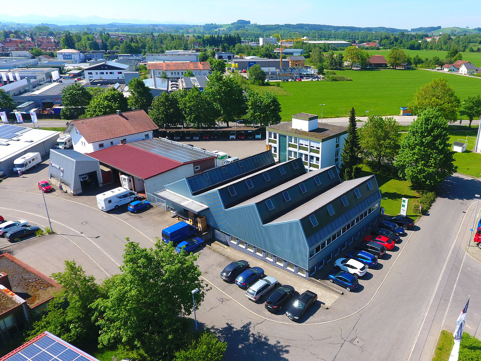 Luftaufnahme eines Industriegebiets mit mehreren Gebäuden, geparkten Autos und Lastwagen, umgeben von grünen Feldern und Bäumen unter klarem Himmel.