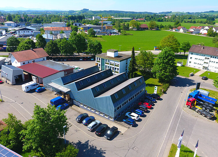 Luftaufnahme eines Industriegebiets mit mehreren Gebäuden, geparkten Autos und Lastwagen, umgeben von grünen Feldern und Bäumen unter klarem Himmel.