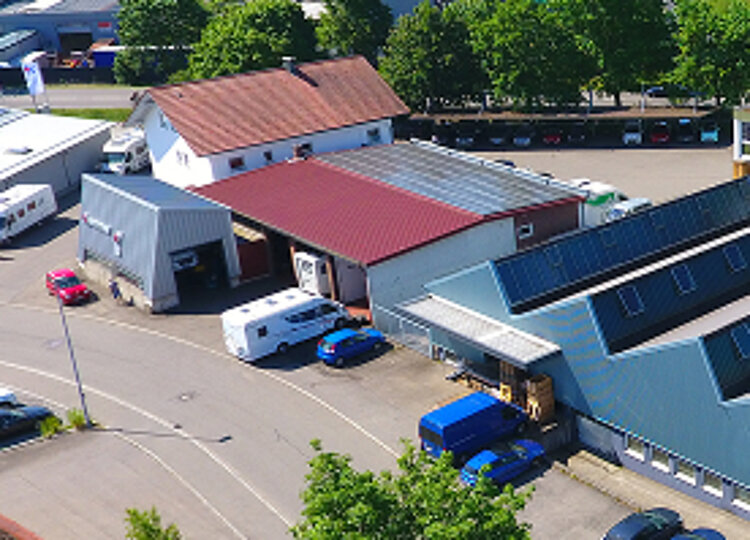 Blick auf ein Gewerbegebiet mit mehreren Hallen, Lagergebäuden und geparkten Fahrzeugen, umgeben von Bäumen und einer Stadtlandschaft im Hintergrund.