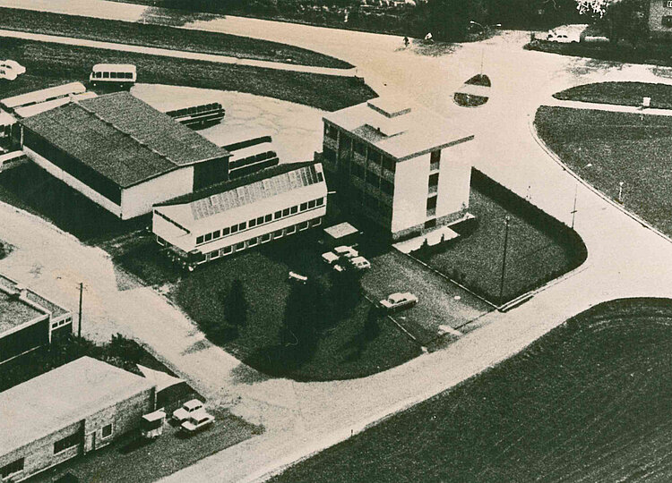 Luftaufnahme eines Gebäudekomplexes mit mehreren Gebäuden, einem Parkplatz mit Fahrzeugen und angrenzenden Straßen in einer ländlichen Umgebung.