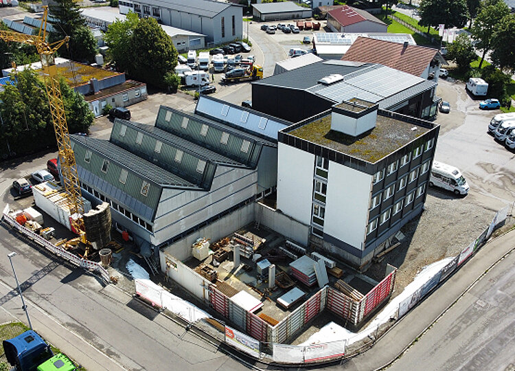 Luftaufnahme eines Industriegebäudes mit Anbau in städtischer Umgebung, umgeben von Bauzäunen und Baumaterialien, daneben eine gelbe Baukran.